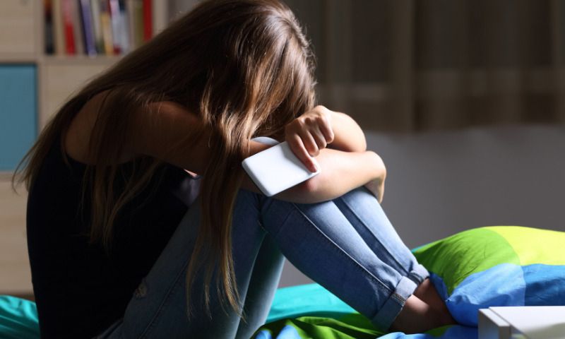 Hilfe bei Mobbing: Junges Mädchen sitzt traurig im Bett mit Smartphone in der Hand