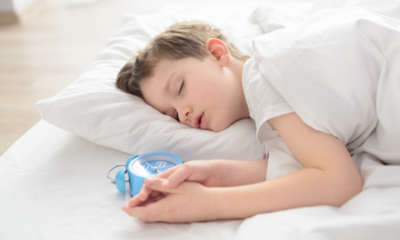 Schlafbedarf von Kinder: Junge schläft im Bett mit Wecker neben sich